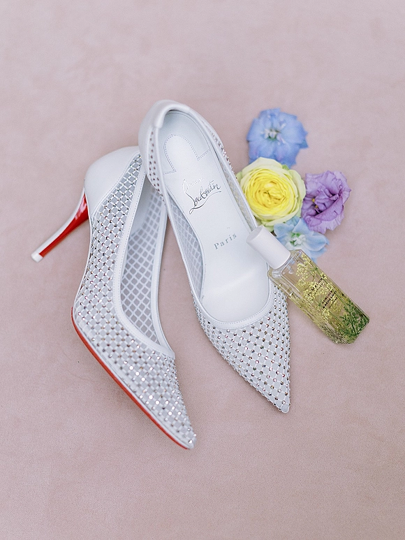 Bridal shoes wedding shoe flatlay of white pointed-toe heels with rhinestone accents, red soles, perfume bottle, and flowers on blush pink surface