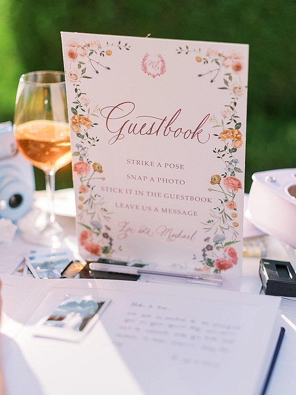 Wedding guestbook table with polaroid guestbook setup, instant camera, photo prints, pens, and a floral sign on a white tablecloth lawn