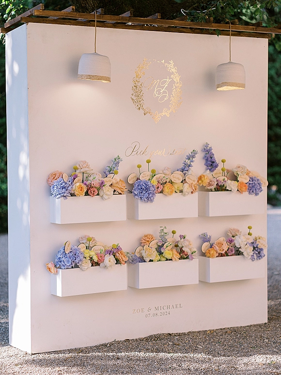 Wedding seating chart on a white wall with gold calligraphy and hydrangea flower boxes under a wood pergola with pendant lights outdoors