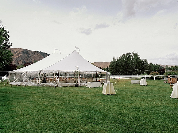 Wedding reception tent with sailcloth canopy and clear sidewalls, cocktail tables and wooden bar set on a lawn by a white fence