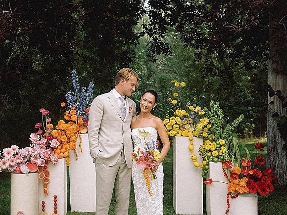 Couple portrait of bride and groom posing on a garden lawn, she holds a colorful bouquet in a strapless lace dress, he wears a light suit with boutonniere, framed by floral plinths with citrus decor