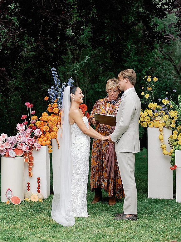 Wedding ceremony moment as bride and groom hold hands while officiant reads vows under floral pillars with citrus accents in garden greenery
