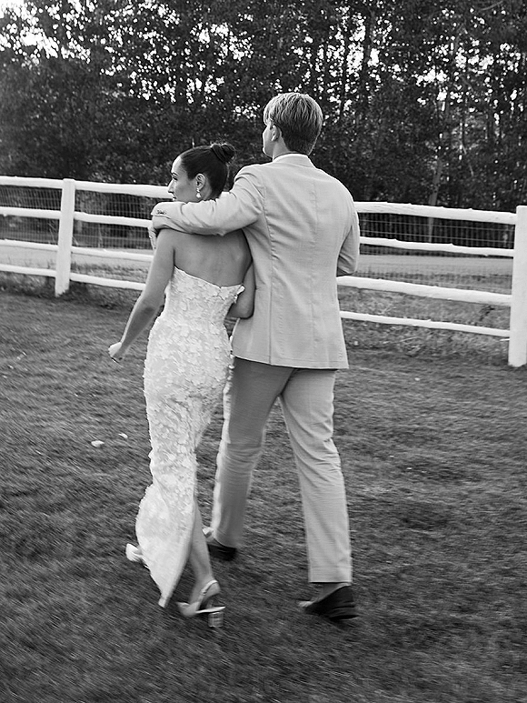 Couple portrait of bride and groom walking away, groom’s arm around her, strapless lace dress, by a white fence in a field