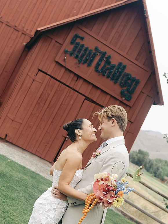 Couple portrait of bride in strapless lace wedding dress and groom in suit embracing with bouquet on lawn before a red barn