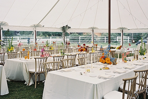 Reception tablescape with colorful wedding centerpieces on white tablecloths, taper candles and place settings under a string-lit clear tent on lawn grass