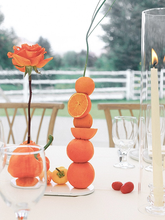 Wedding tablescape with citrus wedding centerpiece of stacked oranges, taper candles in glass hurricanes, wine glasses and place cards on a lawn table