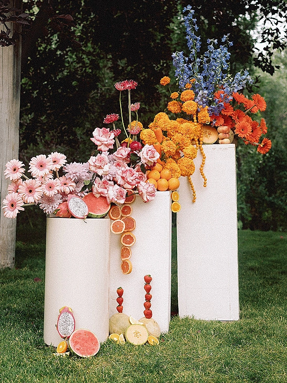 Wedding floral installation with colorful wedding flowers and citrus fruit on white pedestals, set on a garden lawn by a stone wall