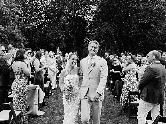 Wedding recessional as bride and groom walking aisle, bride laughing with bouquet and veil while guests applaud on a tree-lined lawn