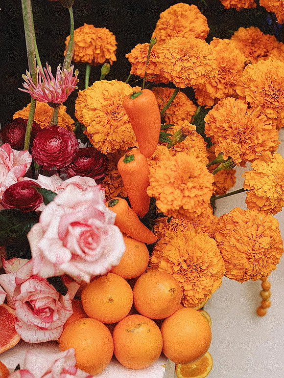 Wedding centerpiece with marigold wedding centerpiece blooms, pink roses and ranunculus, accented by carrots, oranges, and citrus slices on a dark tabletop