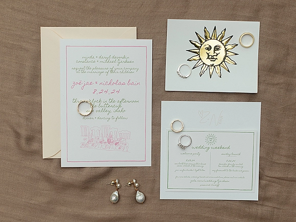 Wedding invitation suite flat lay with envelopes, rings and pearl drop earrings beside an illustrated card on a linen fabric backdrop