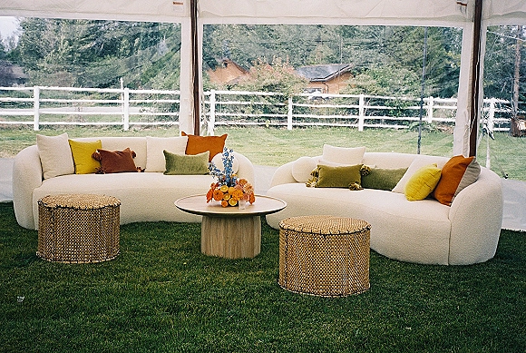 Lounge seating area with wedding lounge furniture, white curved sofas and colorful pillows around a round table under a clear tent on grass lawn