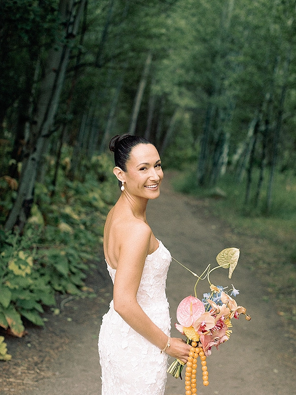 Bridal portrait of a bride looking back on a forest path, holding a tropical bouquet and wearing a strapless lace wedding dress