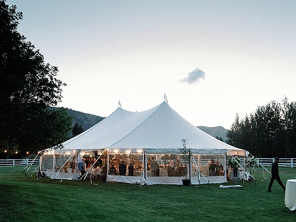 Wedding reception tent with string lights glowing over banquet tables and floral arrangements, set on a lawn with mountain views