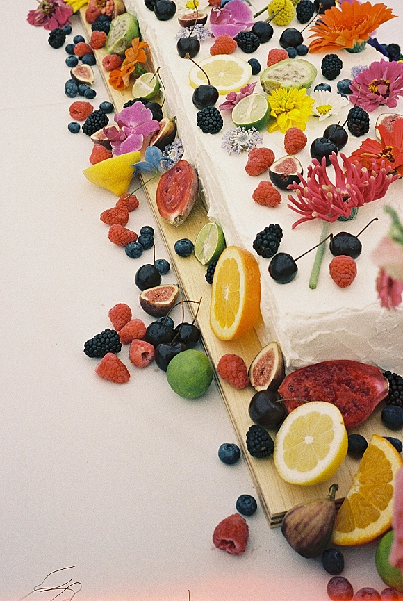 Wedding cake topped with fresh fruit, berries, cherries, figs, and citrus slices on white frosting, set on a white tabletop