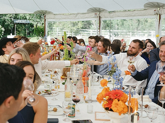 Wedding reception toast as guests raise champagne glasses over a long white table with wildflower centerpiece under a clear tent in daylight
