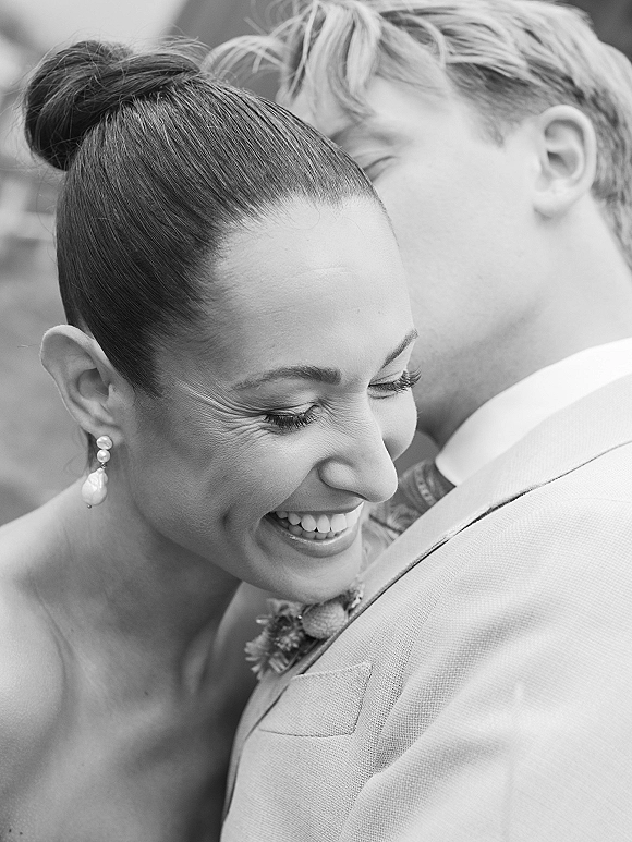 Wedding couple portrait in black and white as the groom whispers to the smiling bride in a close embrace, pearl drop earrings visible outdoors