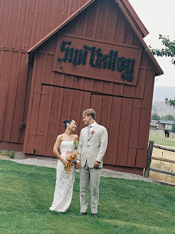 Couple portrait of bride and groom holding hands in front of a red barn, her colorful bridal bouquet against mountains and pasture backdrop