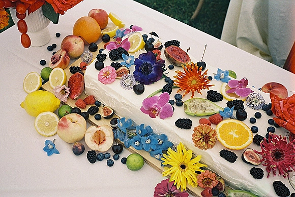 Wedding cake with white buttercream frosting topped with fresh fruit and edible flowers on a wooden board over a white tablecloth outdoors