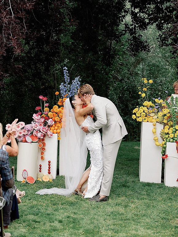 Ceremony kiss at an outdoor wedding ceremony as bride and groom kiss, her veil flowing beside colorful florals and fruit pedestals on a garden lawn