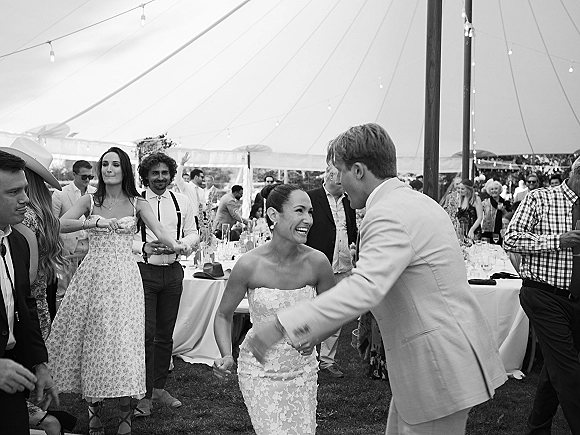 Wedding reception dancing as the bride in a strapless lace gown twirls with groom in a light suit under string lights in a tent crowd