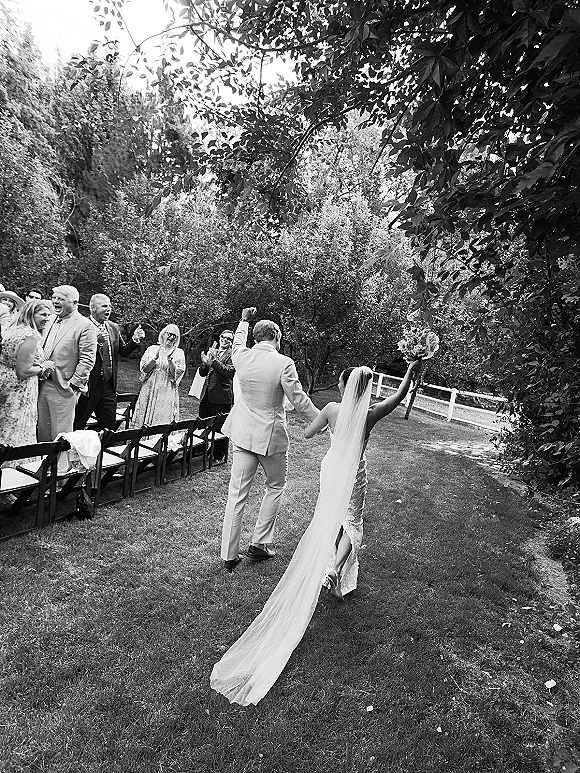 Wedding recessional as newlyweds walking down aisle hand in hand, bride lifting bouquet overhead with long veil, guests by chairs on garden lawn