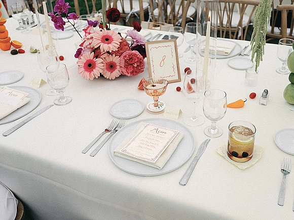 Reception tablescape with a wedding table centerpiece of pink gerbera daisies and orchids, candles, glassware, and framed table number indoors