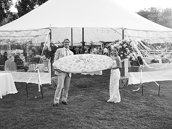 Couple portrait of bride and groom holding a wedding surfboard guest book in front of a clear tent with string lights on a lawn