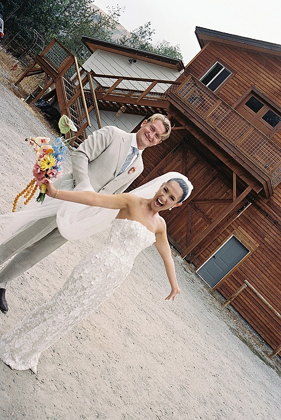 Couple portrait of bride and groom laughing as she holds out a colorful wildflower bouquet, with her veil flowing by a wooden barn