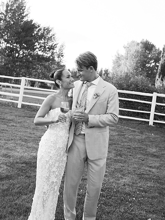 Couple portrait of bride and groom sharing a wedding toast with wine glasses, smiling on a lawn by a white fence and trees