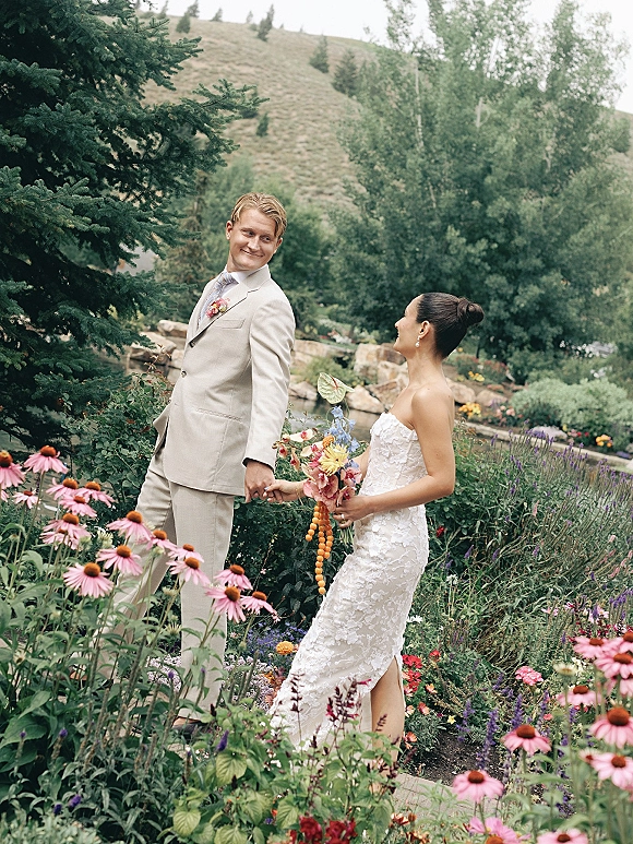 Couple portrait in an outdoor wedding portrait, bride in strapless lace gown holding bouquet, walking hand in hand through gardens
