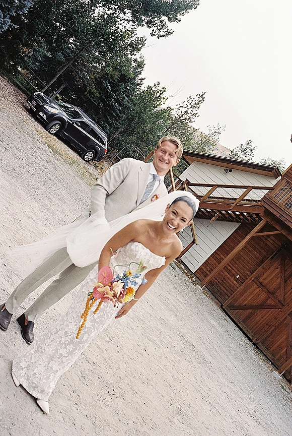 Couple portrait of bride and groom laughing, bride holding a colorful bouquet, walking on a gravel driveway by a wooden building