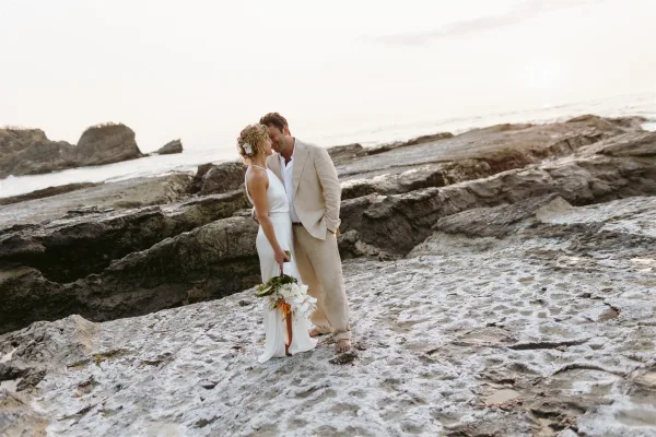 Couple portrait of a beach wedding couple sharing a forehead kiss on coastal rocks by the ocean, bride holding a white orchid bouquet