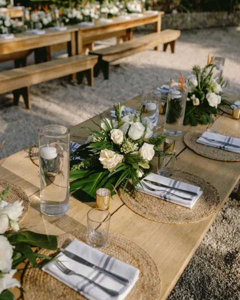 Reception tablescape with a wood farm table wedding setup, white rose and greenery garland centerpieces with candles on a gravel patio in garden greenery