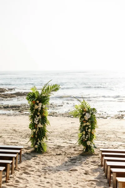Beach ceremony setup with tropical greenery and white floral pillars framing a sandy aisle, wooden chairs facing ocean waves by rocky shore