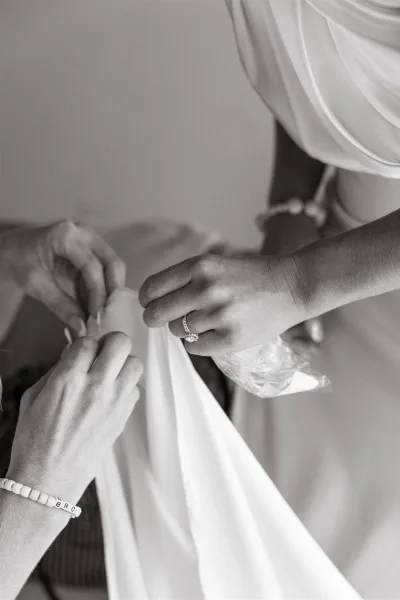Wedding dress fastening as a bridesmaid buttons the bridal gown, showing an engagement ring and bracelet against a plain indoor wall