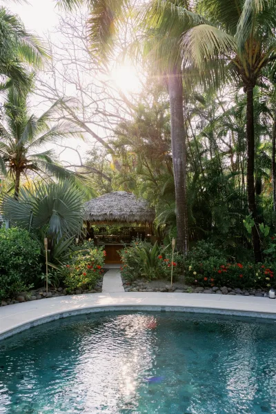 Tropical poolside venue with a thatched roof bar and tiki torches beside a sunlit pool, framed by palm trees and lush foliage
