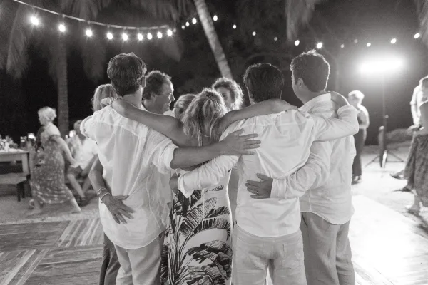 Wedding dance floor packed with wedding guests dancing in a tight group hug under string lights, with palm trees and stage lighting at night