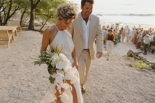 Ceremony recessional as bride and groom walking barefoot on the beach, bride holding a white orchid bouquet, guests by the shoreline