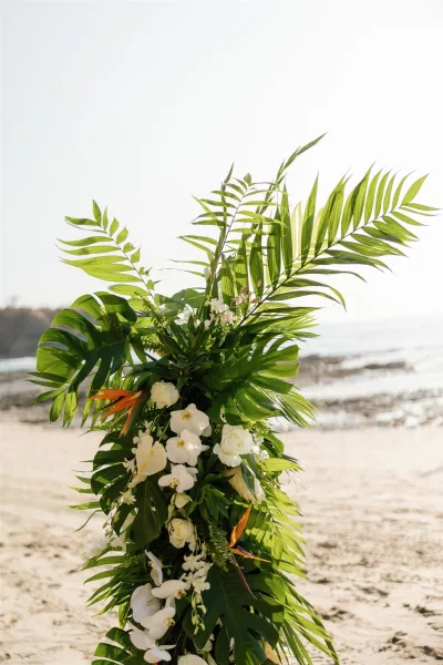 Wedding ceremony florals with tropical wedding altar flowers, palm fronds and white orchids on a stand by the sandy ocean shoreline