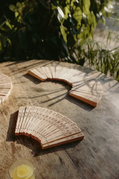 Wedding hand fans arranged on a rustic wooden table beside a cocktail glass, with greenery and soft outdoor light in the background