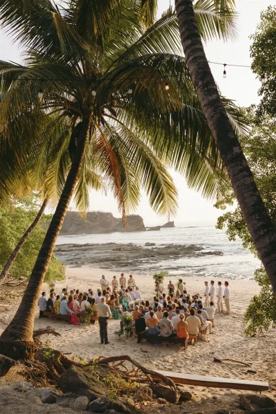 Beach wedding ceremony with destination wedding ceremony setup on sand, wood benches and greenery aisle markers facing the ocean and palms