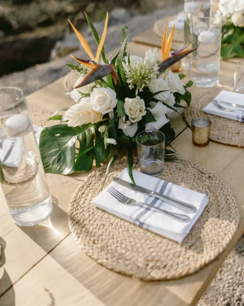 Reception tablescape with tropical wedding centerpiece featuring bird of paradise and white roses, monstera leaves, woven placemats, amber votives on a wood table outdoors