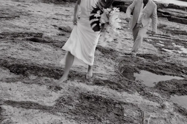 Wedding couple portrait of a bride and groom running barefoot along a rocky beach, bride in white dress holding a bouquet by tide pools