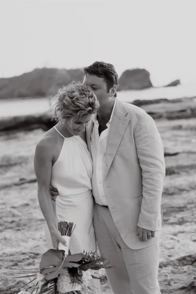 Couple portrait of a beach wedding couple sharing a forehead kiss, bride holding a bouquet on a rocky ocean coastline with cliffs beyond
