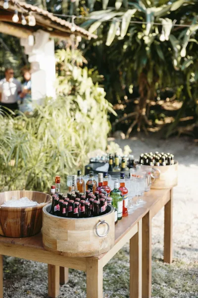 Wedding bar setup with outdoor wedding bar on a wood table, ice buckets, beer and wine bottles, glassware and string lights amid tropical greenery