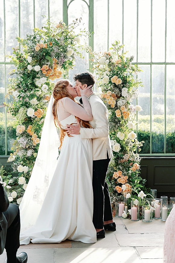 Wedding kiss as bride in a strapless dress and veil meets groom in a cream tuxedo under a rose arch with candles in a greenhouse