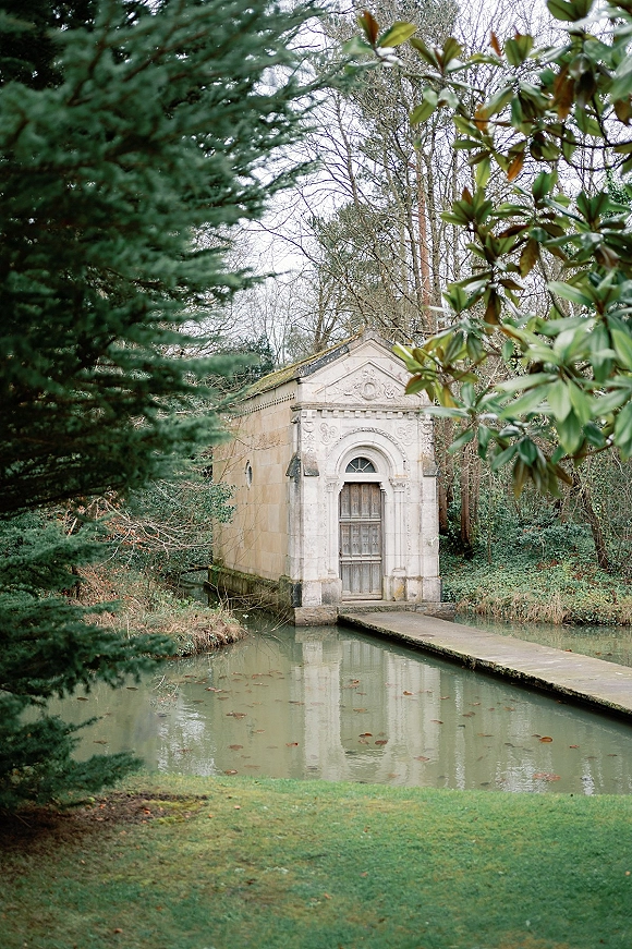 Wedding venue exterior with a stone building and arched doorway, wooden door, and stone walkway beside a pond under overcast trees