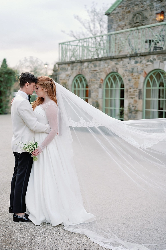 Wedding couple portrait with bride and groom forehead touch, her cathedral veil trailing as they embrace in a stone courtyard backdrop