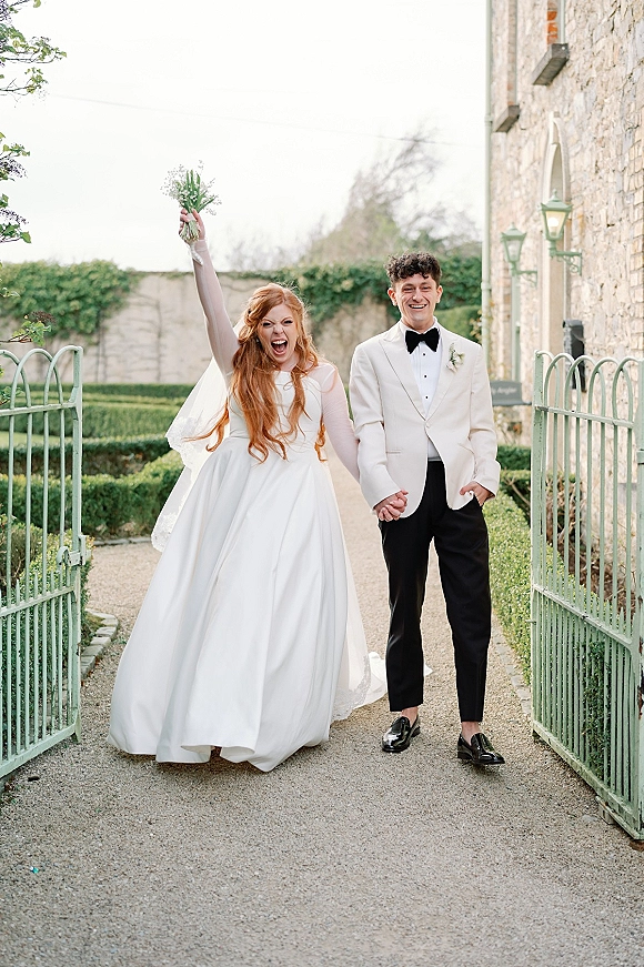 Couple portrait of bride and groom holding hands as she raises her bouquet, long veil trailing by wrought iron gates on a garden path