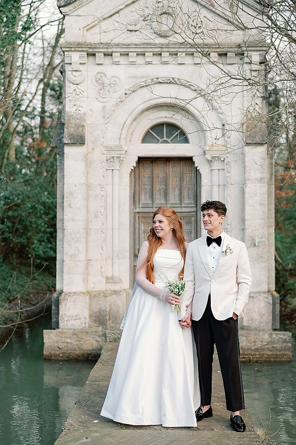 Couple portrait of bride and groom holding hands, smiling outside a stone chapel, bride in satin gown with lily of the valley bouquet